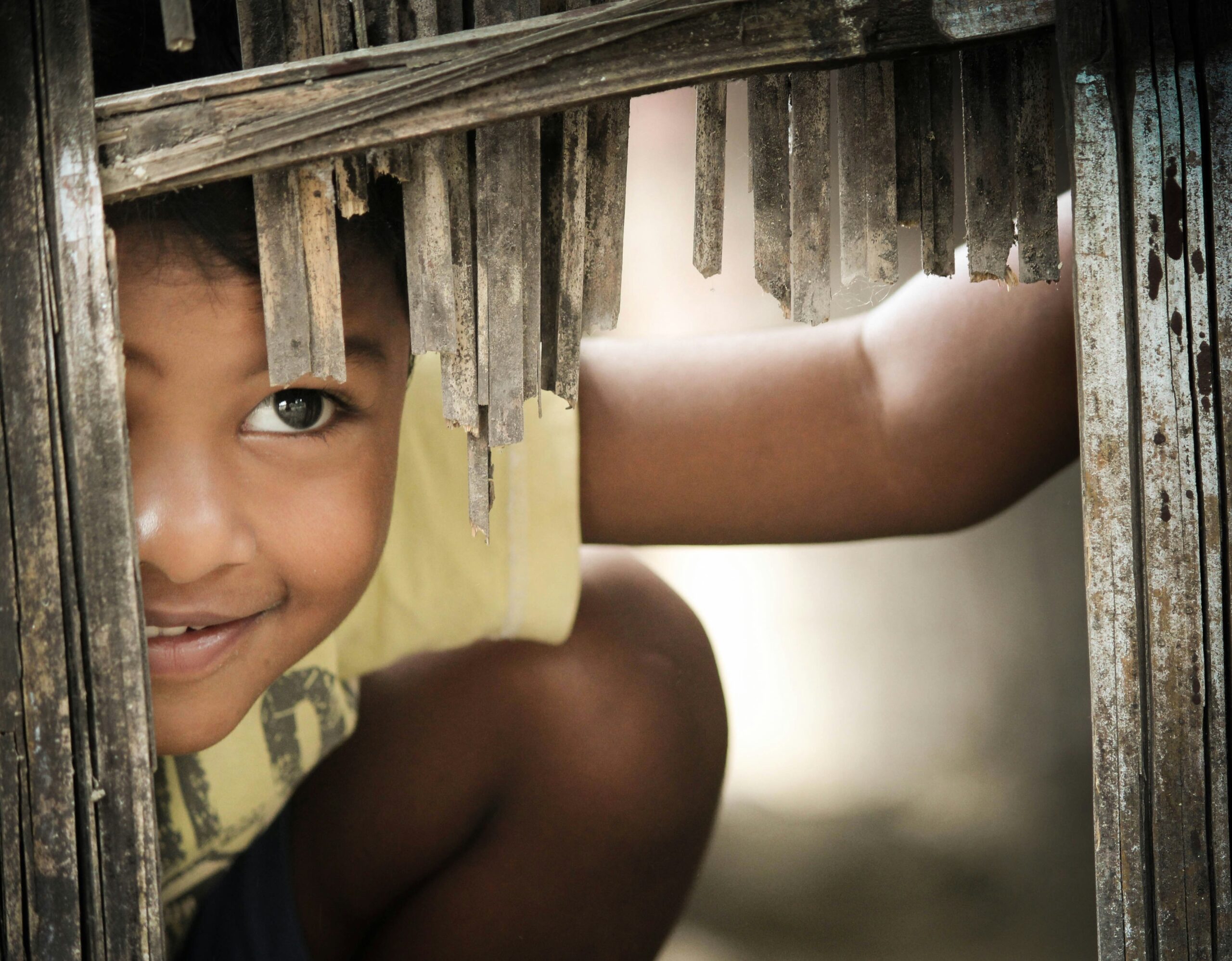 A young boy smiling cheekily through a gap in a fence.